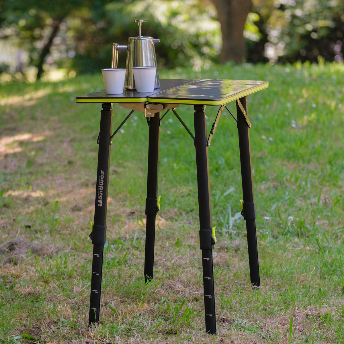 A portable folding camping table with a black and yellow top and black legs, set outdoors on a grassy lawn. A stainless steel coffee pot and two white cups are placed on the tabletop.